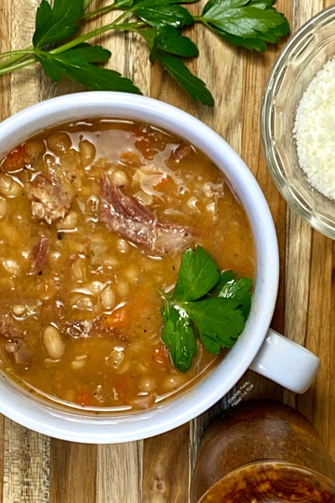 Instant Pot Ham and Bean Soup in a white bowl with handles. The bowl sits atop a wooden cutting board with a sprig of parsley and a ramekin of grated Romano cheese.