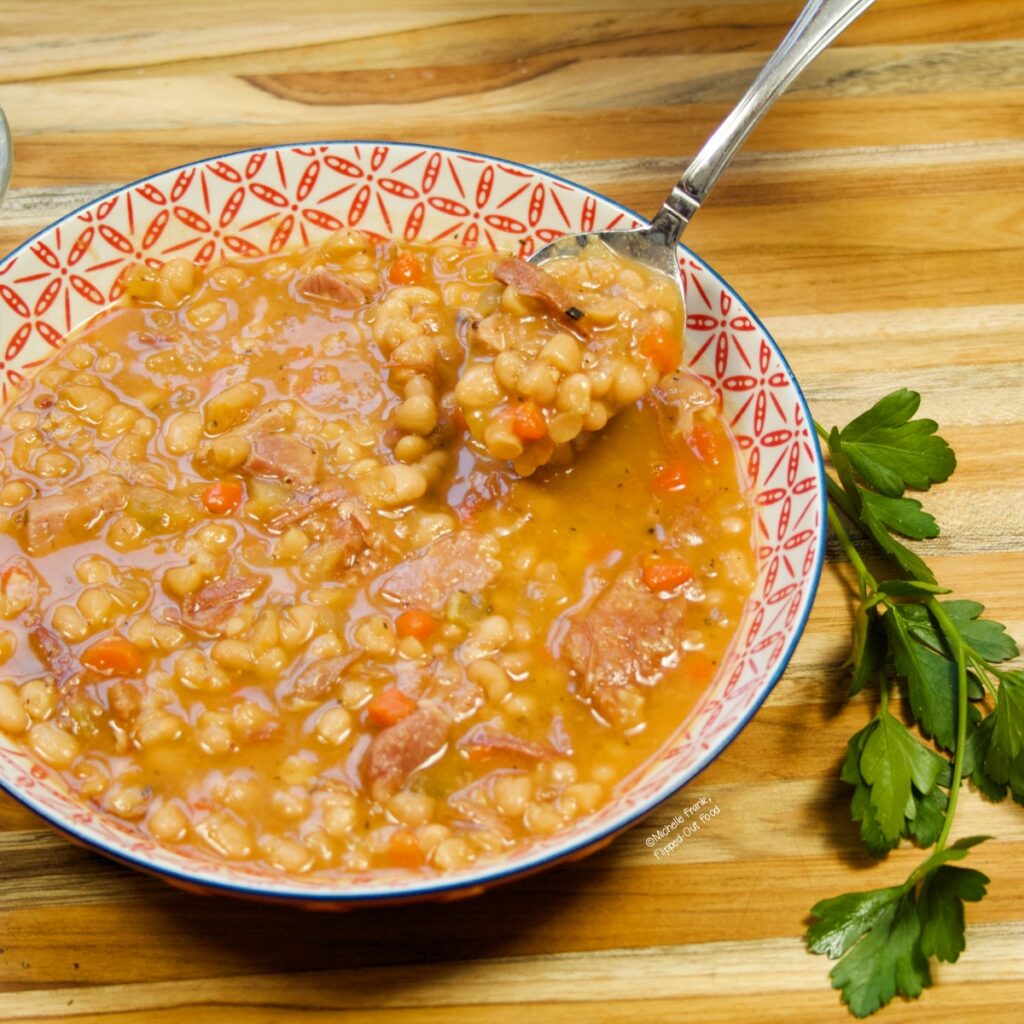 A red and white bowl with a serving of Instant Pot Ham and Bean Soup. A spoonful of the soup is being raised toward the camera.