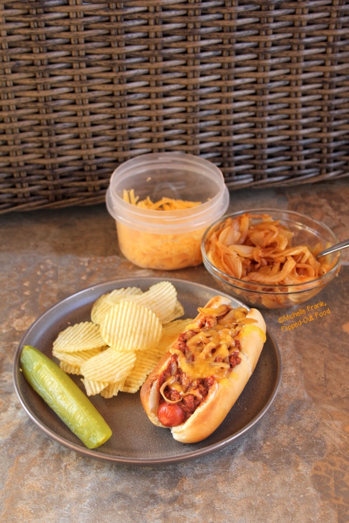 Side view of a chili cheese dog on a plate with wavy potato chips and a pickle spear. Behind the plate are a bowl of caramelized onions and a plastic container with grated cheese.