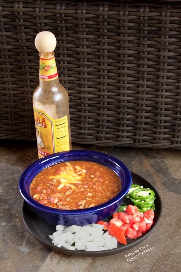 Quick, Easy Turkey Chili topped with Colby-Jack cheese in a blue bowl on top of a plate with chopped onions and tomatoes and sliced jalapenos. A bottle of Cholula sits in the background.