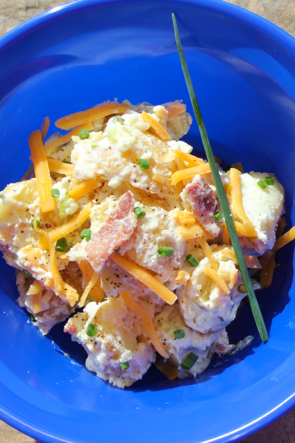 A blue bowl with a serving of loaded baked potato salad garnished with sliced chives and a single whole chive.