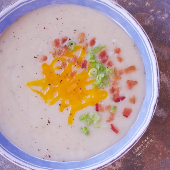 Pureed Cauliflower Loaded Baked Potato Soup: a closeup of a serving in a blue and white bowl, topped with cheddar cheese, bacon, and sliced scallions.