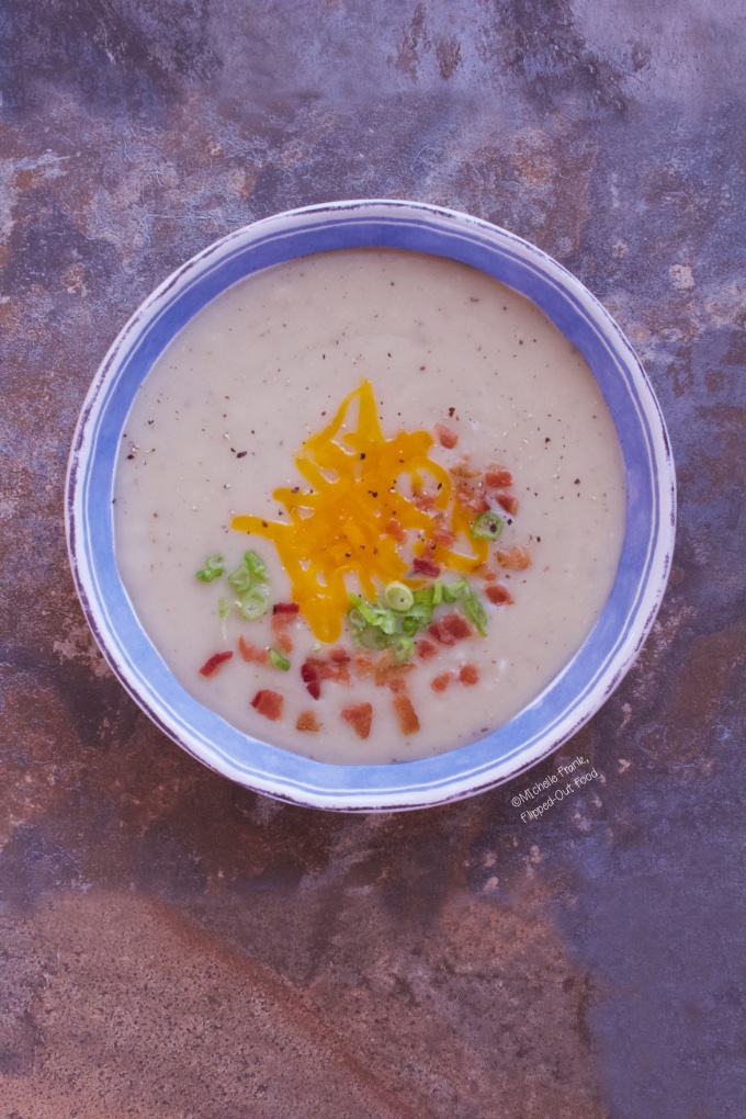 Pureed Cauliflower Loaded Baked Potato Soup: a serving in a blue and white bowl, topped with cheddar cheese, bacon, and sliced scallions.