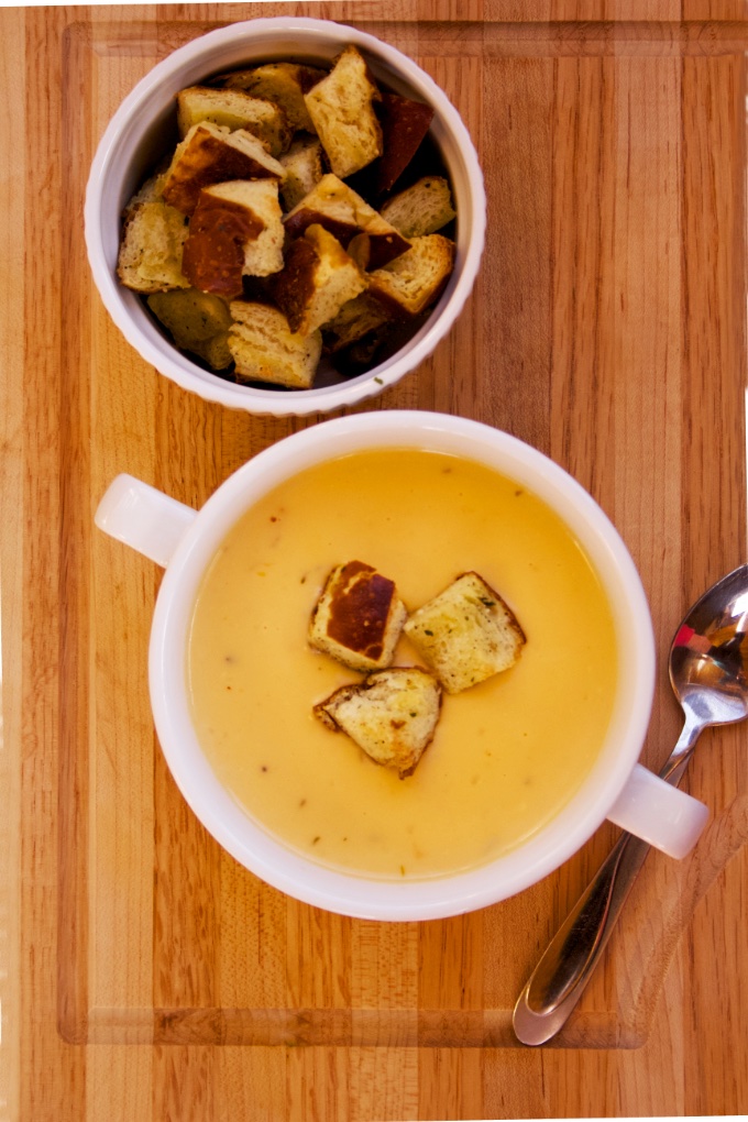 Beer Cheese Soup with Pretzel Croutons: a serving of the soup in a white bowl with handles. 3 croutons float on the soup and a ramekin with additional croutons sits behind the bowl.