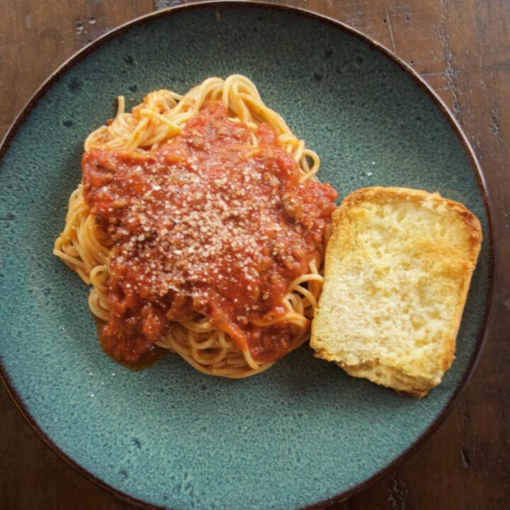 Easy Cheat Bolognese Sauce on spaghetti, served on a blue stoneware plate with a piece of garlic bread. Crumbled Romano cheese is sprinkled over the top.