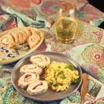 Side view of a plate with four slices of work-ahead chicken rollatini next to a serving of lemon-broccoli orzotto. In the background, a platter with slices of work-ahead chicken rollatini sits next to a glass of white wine.
