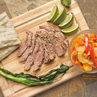Top view of carne asada with fajita vegetables arranged on a cutting board with a folded flour tortilla, grilled scallions, lime wedges, and the cut end of a jalapeno pepper.