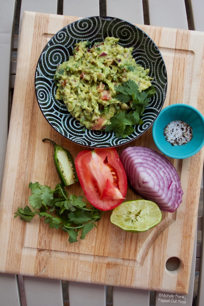 Guacamole in a black bowl set atop a wooden cutting board. Next to the bowl sits a ramekin with salt and pepper, a cut red onion, squeezed lime, cut tomato, cut jalapeno, and a sprig of cilantro.