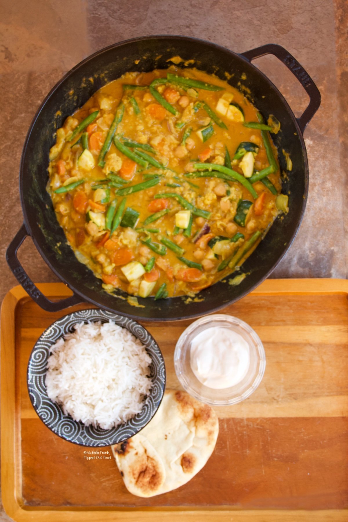 Easy Red Vegetable Curry in a cast-iron wok next to a bowl of rice, a pita, and a ramekin of Greek yogurt.