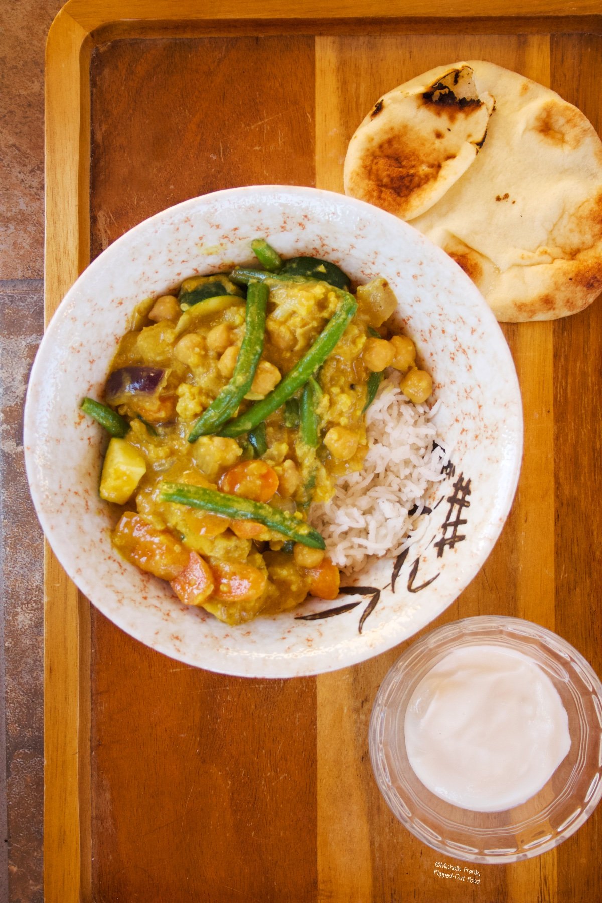 Easy Red Vegetable Curry in a ceramic bowl over rice. The bowl sits next to a pita and a ramekin of Greek yogurt atop a wooden serving tray.