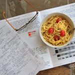 A serving of Healthy MIcrowave Dorm Room Pasta with sliced grape tomatoes. The bowl sits on top of a lab notebook next to an unoccupied pair of glasses.