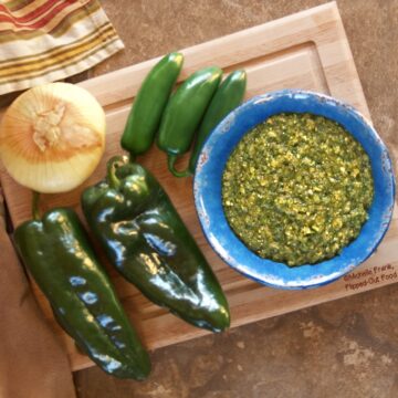 A rotated view of fresh salsa verde in a blue bowl sitting atop a wooden cutting board, surrounded by poblano and jalapeno peppers and an onion. A colorful, striped cloth sits off to one side.
