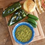 Overhead view of fresh salsa verde in a blue bowl sitting atop a wooden cutting board. The bowl is surrounded by two poblano peppers, three jalapenos, and an onion.