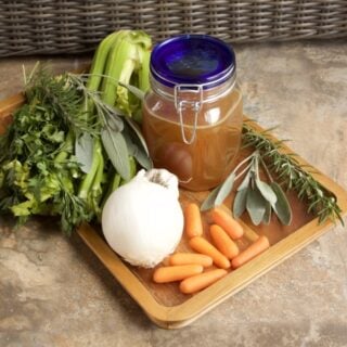 Crockpot Turkey Stock in a blue-capped Mason jar. Surrounded by herbs, carrots, onion, garlic, and celery.