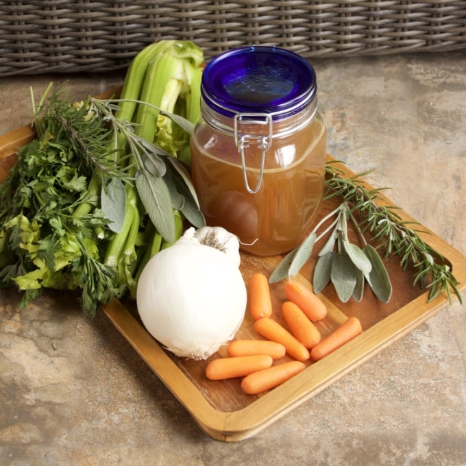 Crockpot Turkey Stock in a blue-capped Mason jar. Surrounded by herbs, carrots, onion, garlic, and celery.