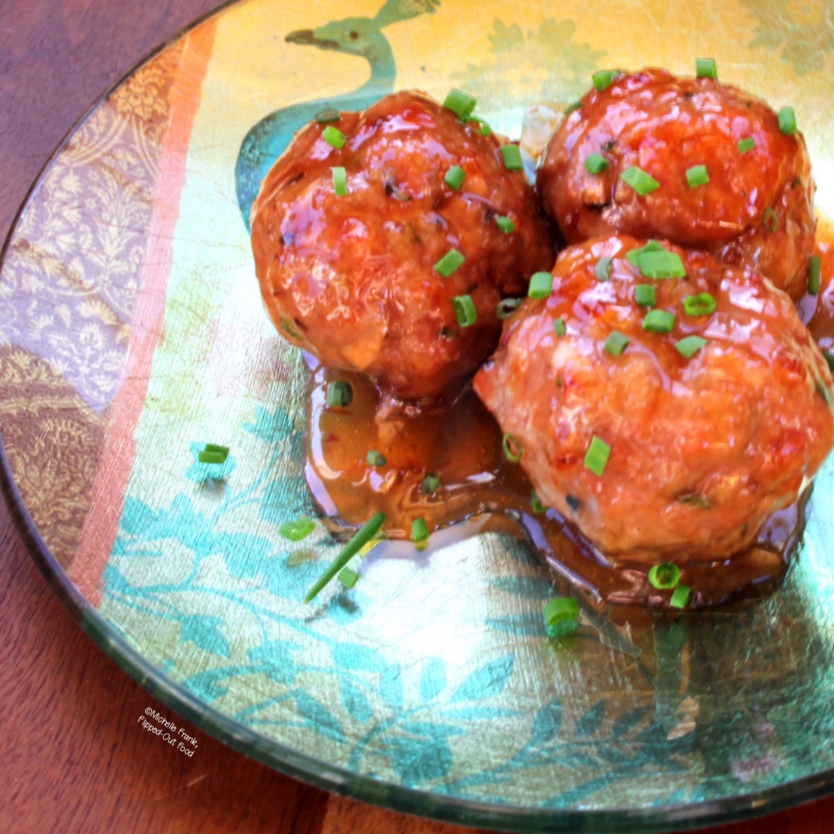 Three sweet & zingy Asian meatballs on a decorative party plate. The meatballs are garnished with minced chives.