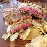 Side view of Best-Ever Reuben Sandwiches served with 1000 island dressing on the side in a clear ramekin and a serving of chips. #reubensandwich #saintpatricksday #stpaddysday #cornedbeef #pubfood #sandwich via @flippedoutfood