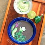 Slow-Cooker Black Bean Soup, in a blue bowl with avocado, cilantro, jalapenos, red onion, and avocado crema with lime and jalapeno in the background. #blackbeansoup #mexicanfood #slowcooker #crockpot #soup @FlippedOutFood