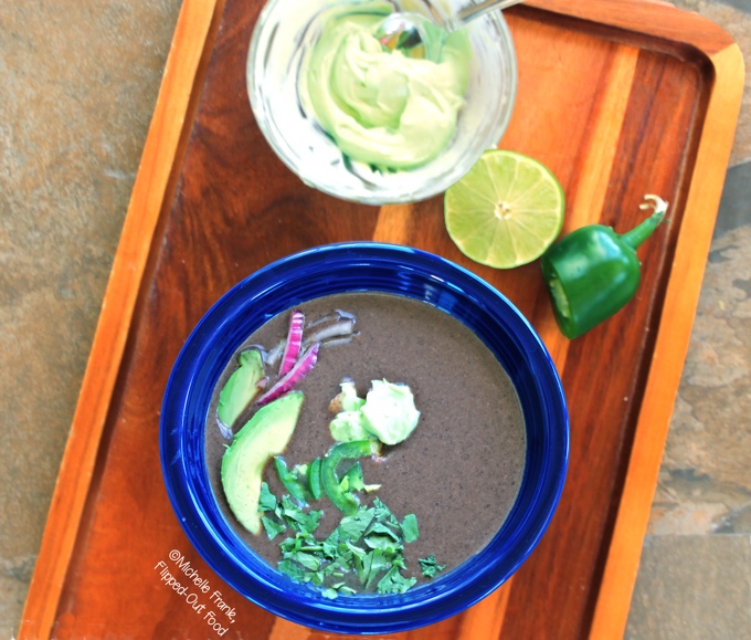 Slow-Cooker Black Bean Soup, in a blue bowl with avocado, cilantro, jalapenos, red onion, and avocado crema with lime and jalapeno in the background. #blackbeansoup #mexicanfood #slowcooker #crockpot #soup @FlippedOutFood