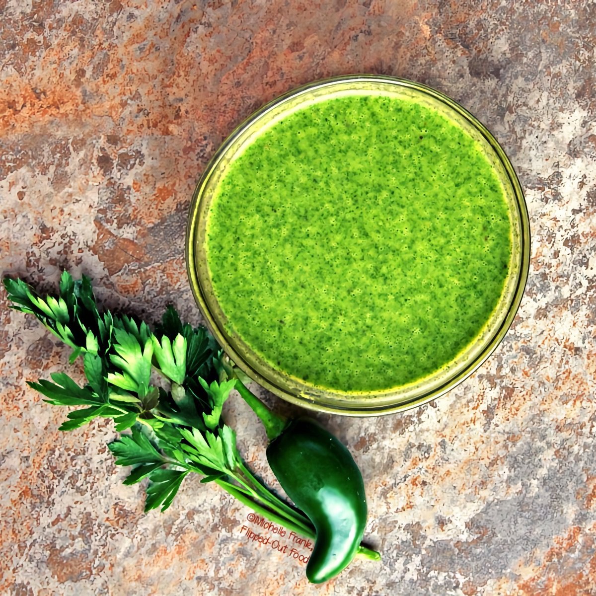 An overhead view of spicy chimichurri sauce, blended smooth and served in a ramekin. A jalapeño pepper and a sprig of parsley sit nearby.