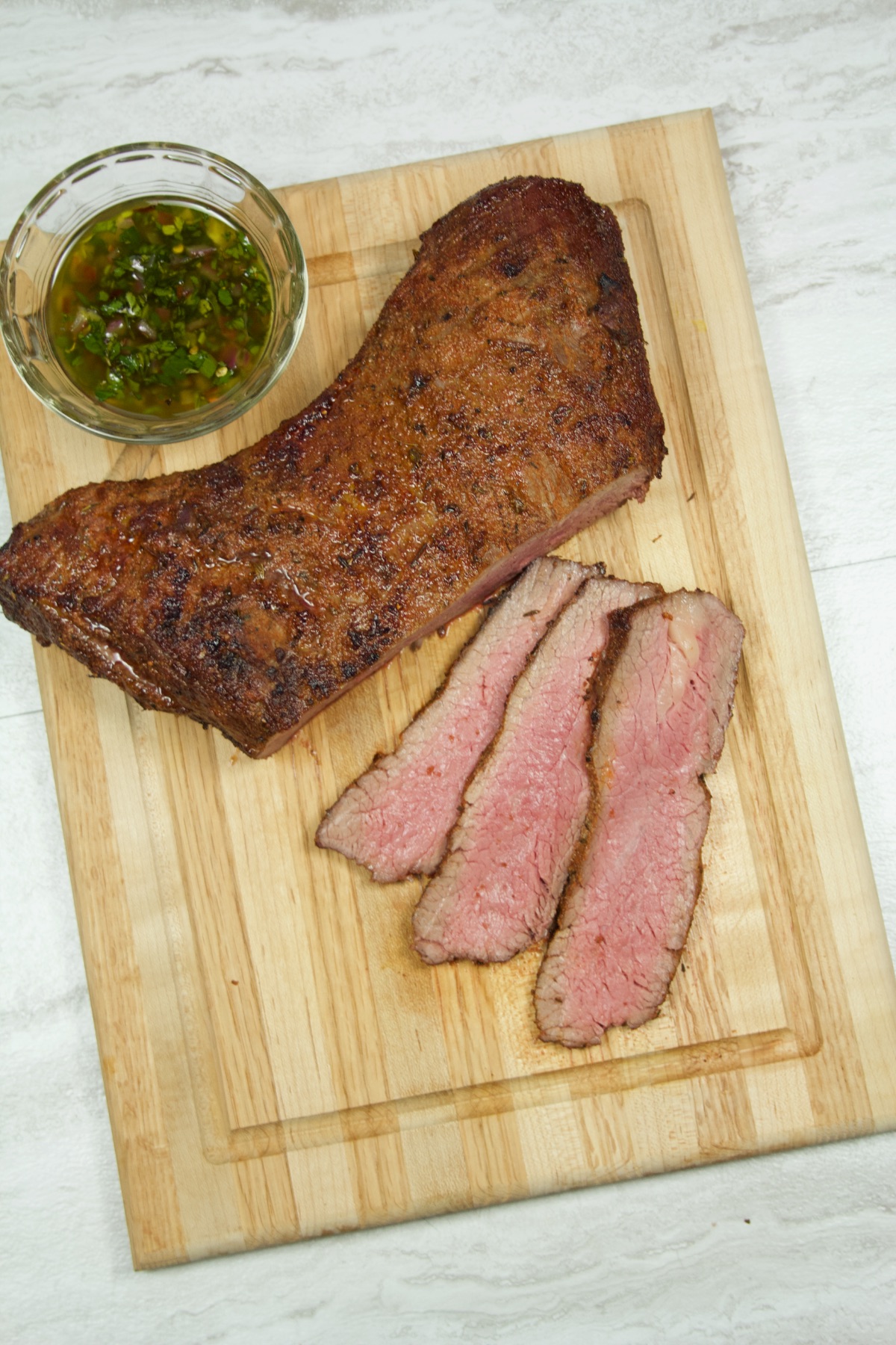 Overhead view of a tri-tip roast cut into thin steaks, with a ramekin of chimichurri sauce sitting nearby.
