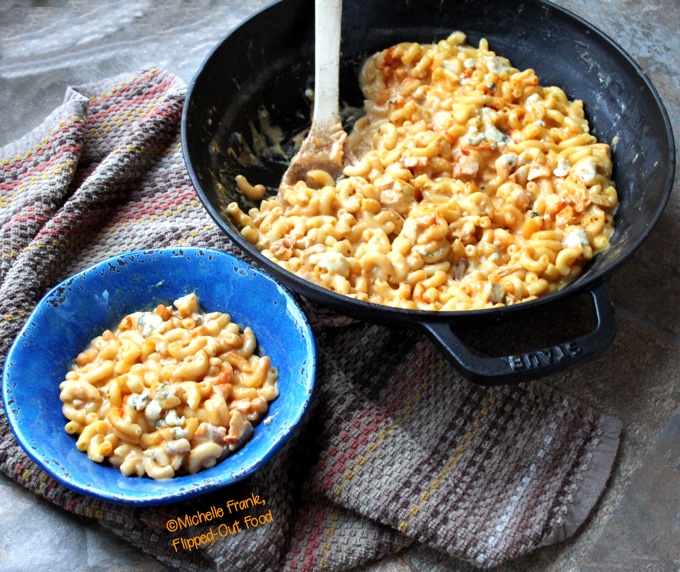 One-Pot Buffalo Chicken Macaroni and Cheese: side view showing a vivid blue bowl with mac and cheese sits in the foreground in front of a cast-iron skillet full of the Buffalo chicken macaroni and cheese.