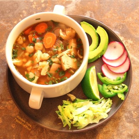 Green Chile Chicken Posole (Posole Verde de Pollo): a side view of a white, 2-handled bowl full of the posole sitting on a plate piled with garnishes, including avocado, thinly sliced radishes, jalapeno, lime, and shredded lettuce.