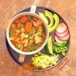 Green Chile Chicken Pozole (Pozole Verde de Pollo): a top view of a 2-handled bowl full of the pozole sitting on a plate piled with garnishes, including avocado, thinly sliced radishes, jalapeno, lime, and shredded lettuce.