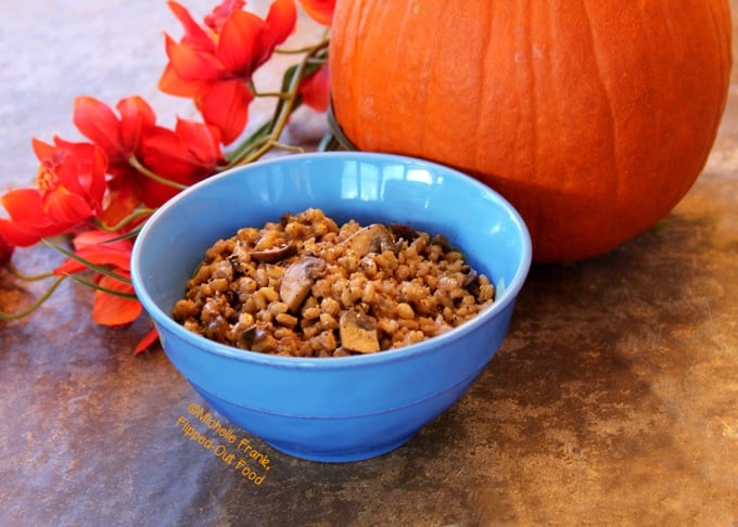 Side view of a blue bowl filled with double-mushroom barley pilaf. The bowl is sitting in front of a pumpkin and a bunch of red flowers.