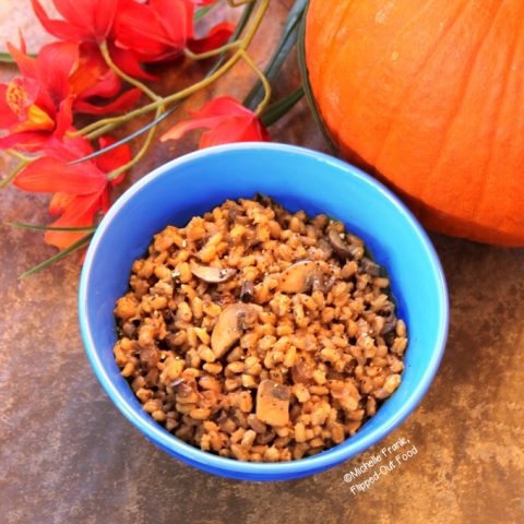 Top view of a blue bowl filled with double-mushroom barley pilaf. The bowl is sitting in front of a pumpkin and a bunch of red flowers.