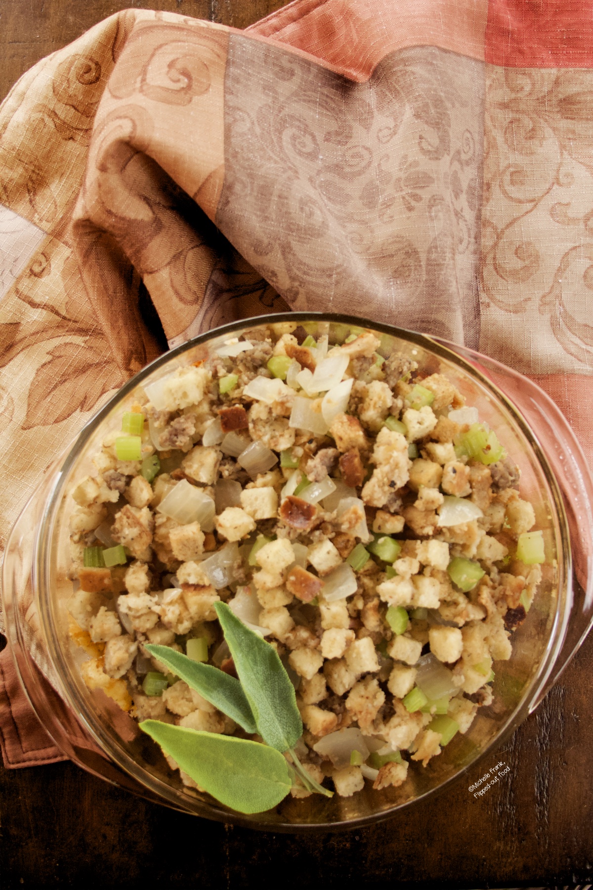 A round Pyrex casserole dish of Easy Sausage Stuffing with a garnish of sage, set atop a colorful cloth.