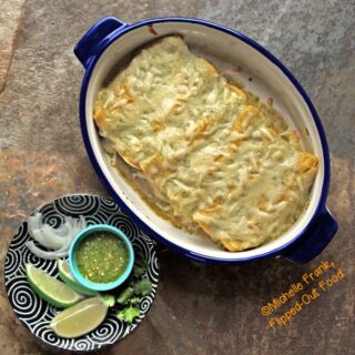 A blue casserole dish full of creamy green chile turkey enchiladas, next to a decorative plate of garnishes, including lime wedges, sliced white onions, cilantro, and a ramekin of salsa verde.