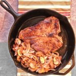Steak Mushroom Onion Skillet: a porterhouse steak in a cast-iron skillet with caramelized mushrooms and onions. The skillet sits atop a colorful striped cloth.