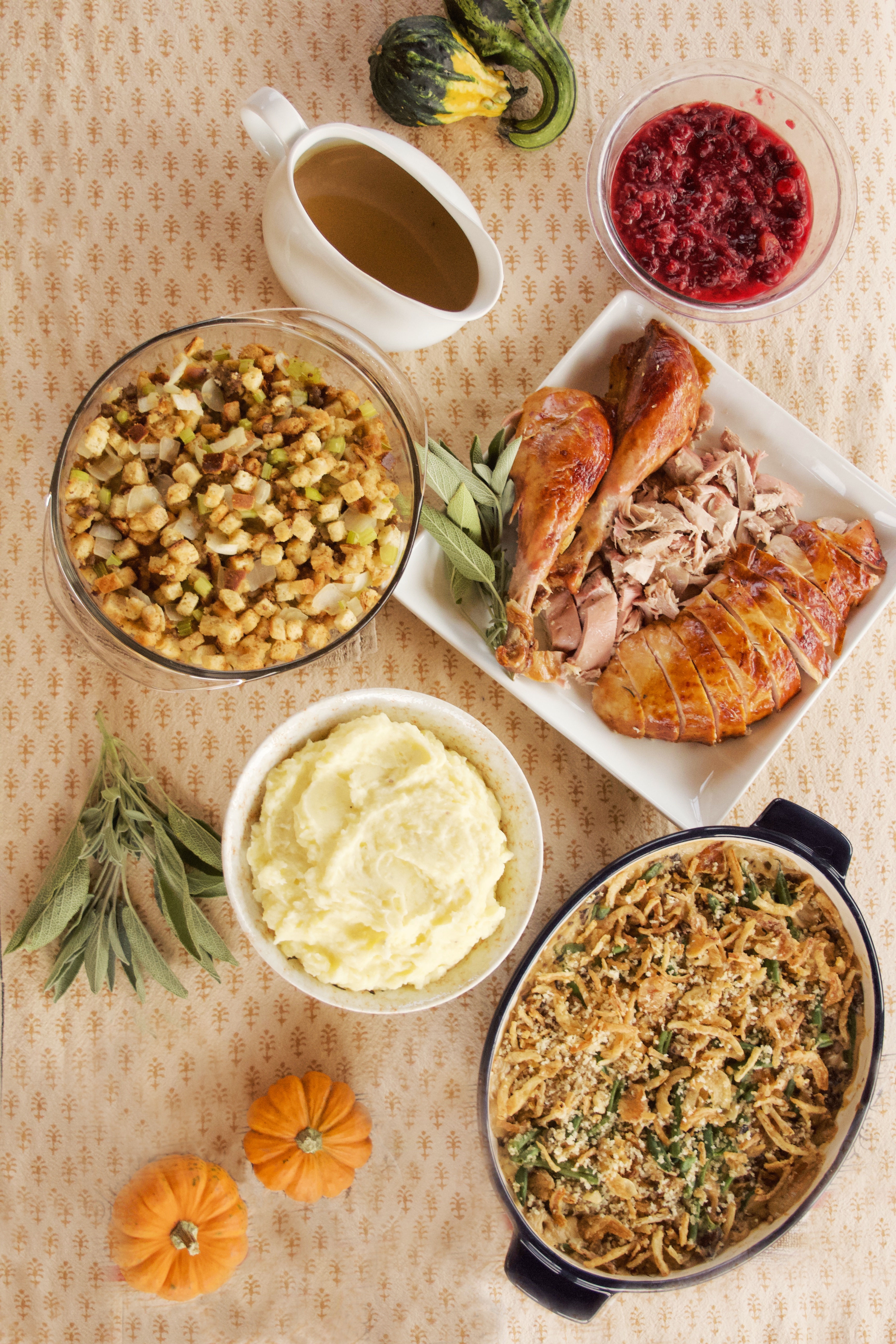 The Complete Work-Ahead Holiday Dinner Meal Plan spread (clockwise, from top left): Easy, Rich Turkey Gravy, Orange-Ginger-Spice Cranberry Sauce, Ultimate Classic Roast Turkey, Make-Ahead Green Bean Casserole, Creamy Meal-Prep Mashed Potatoes, and Easy Sausage Stuffing. The spread is accented with tiny gourds and a sprig of sage.