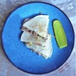 Three wedges of Cubano Quesadillas arranged on a bright blue plate next to a slice of dill pickle.