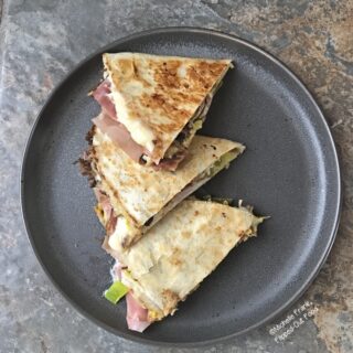 Three wedges of Cubano Quesadillas arranged on a dark grey plate next to a slice of dill pickle.