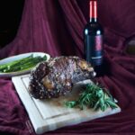 Date Night Prime Rib roast resting on a cutting board surrounded by herbs (rosemary and sage). In the background is a dish of roasted asparagus and a bottle of red wine.