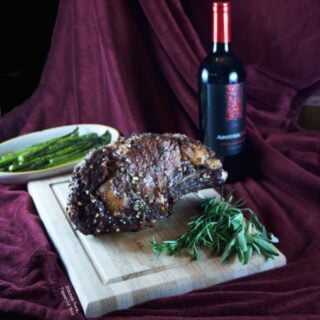 Date Night Prime Rib roast resting on a cutting board surrounded by herbs (rosemary and sage). In the background is a dish of roasted asparagus and a bottle of red wine.