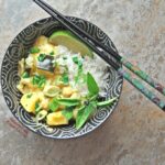 A top view of Easy Squash Curry in a decorative black bowl with white swirls. A pair of ornate chopsticks sits on top. The curry is served over basmati rice, with a sprig of Thai basil and a lime wedge.