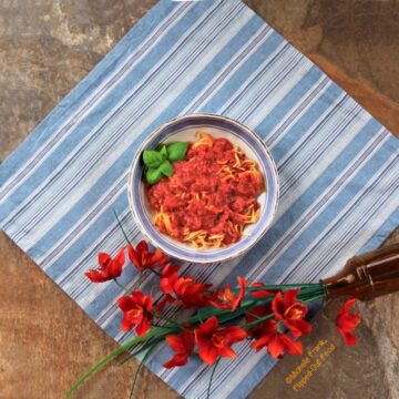 Top view of a bowl of chunky pantry marinara sauce in a light blue and white bowl sitting atop a blue and white striped cloth. The bowl sits next to a bunch of bright orange flowers. The spaghetti is garnished with a sprig of basil.