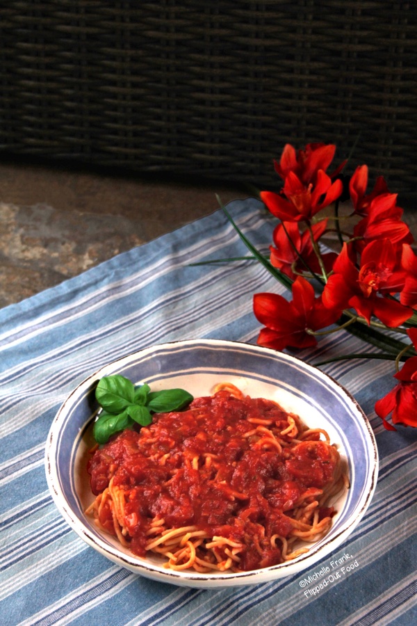 Side view of a serving of chunky pantry marinara sauce in a light blue and white bowl sitting atop a blue and white striped cloth. The bowl sits next to a bunch of bright orange flowers. The spaghetti is garnished with a sprig of basil.