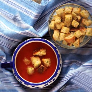 Top view of a serving of Vegan Pantry Tomato Soup with Homemade Croutons in a decorative blue and white, single-handled bowl. The bowl sits on a blue and white-striped cloth. A bowl full of more croutons sits just behind and to the right.