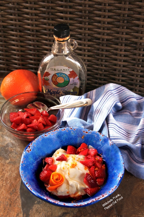 Side view of Maple-Orange-Strawberry Yogurt in a bright blue bowl. A piece of orange zest has been arranged to look like a flower. The bowl sits in front of a blue-and-white-striped cloth, a bowl with more strawberries, an orange, and a jug of maple syrup.