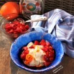 Side view of Maple-Orange-Strawberry Yogurt in a bright blue bowl. A piece of orange zest has been arranged to look like a flower. The bowl sits in front of a blue-and-white-striped cloth, a bowl with more strawberries, an orange, and a jug of maple syrup.