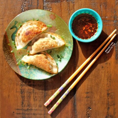 Top view of Pork-Shrimp Wontons with Soy-Ginger Dipping Sauce. Three wontons are arranged on a decorative, iridescent appetizer plate sprinkled with chopped chives. A blue ramekin with dipping sauce sits next to the plate and an ornate pair of chopsticks.