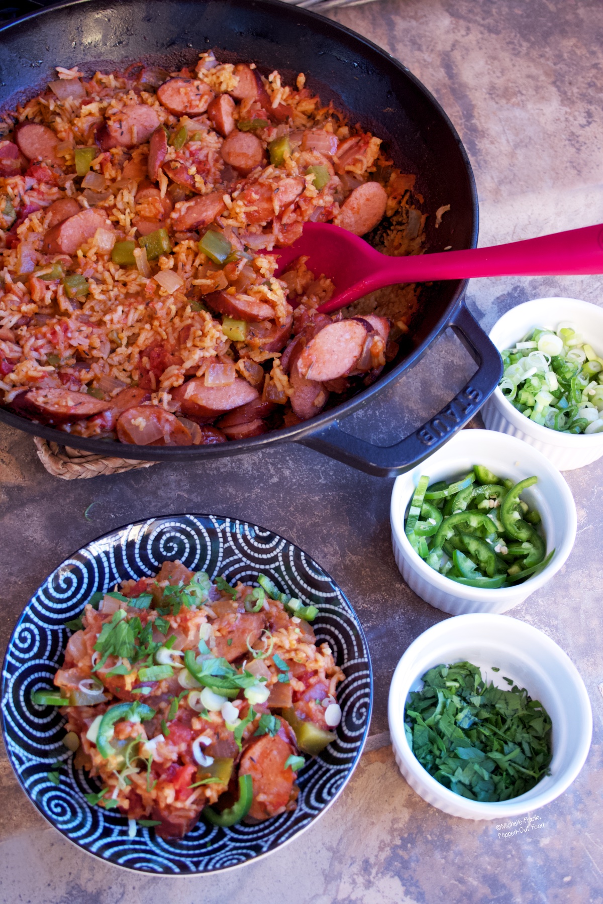 Cajun Sausage Rice Skillet with a serving dished out into a bowl in the foreground. Ramekins with scallions, jalapenos, and parsley sit to the side.
