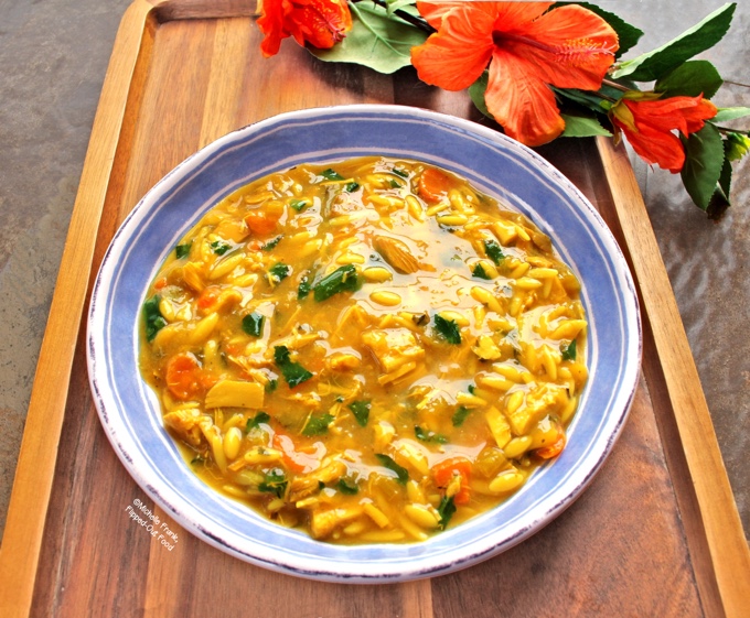 A serving of lemon-turmeric chicken-orzo soup in a blue and white bowl sitting on a wooden tray with hibiscus flowers in the background. The soup is full of baby kale, chicken, carrots, and onions. The turmeric gives the soup a bright yellow color.