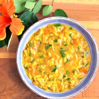 A serving of lemon-turmeric chicken-orzo soup in a blue and white bowl sitting on a wooden tray with hibiscus flowers in the background. The soup is full of baby kale, chicken, carrots, and onions. The turmeric gives the soup a bright yellow color.