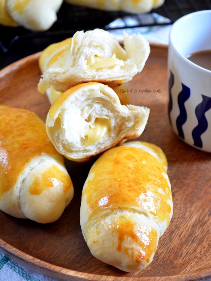 4 Asian-Inspired Dishes, #1. Filipino Cheese Bread Rolls: two whole rolls on a wooden platter next to a cup of coffee. One roll that has been torn in half sits on top, showing the melty goodness of the cheese inside.