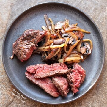 Top view of a round, grey plate with sliced ribeye steak with caramelized onions and mushrooms. The steak is cooked to a perfect rare doneness.